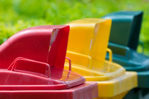 A row of three large plastic wheelie bins used for rubbish collection, positioned on a grassy outdoor area near a pavement or driveway. The first bin in the foreground is red with a textured surface and a sturdy handle, followed by a yellow bin with a slightly worn finish, and a dark green bin at the end, all with hinged lids. The bins are arranged side by side, slightly angled, with the red and yellow bins slightly overlapping each other. The environment appears to be a residential or commercial property exterior, with a blurred background of green grass and possibly some trees, suggesting a typical waste disposal setup. The scene is well-lit with natural daylight, emphasizing the vibrant colours of the bins and the textures of the plastic material. This image relates to rubbish removal services, such as those provided by Commercial Waste Yiewsley, serving local businesses and residents in the Yiewsley area, which is part of the broader London boroughs near Postcode WD or UB.