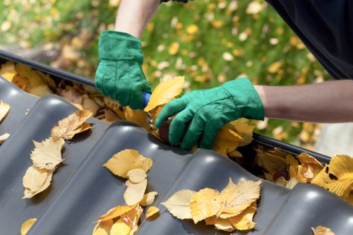 A person wearing green gloves is collecting fallen yellow and brown autumn leaves and placing them into a grey plastic gutter with a corrugated surface, which is situated outdoors amid a background of green foliage and scattered leaves on the ground. The gutter is attached to a building or structure and is being cleared of debris to prevent blockage, in a typical domestic or commercial environment in Yiewsley. The image emphasizes outdoor maintenance work related to rubbish and waste removal, with the focus on manual leaf clearing and the textured surface of the gutter. Light and shadow suggest natural daylight, and the scene reflects routine seasonal cleaning activities often undertaken by commercial waste removal services in the local area or surrounding boroughs under the domain of Commercial Waste Yiewsley.