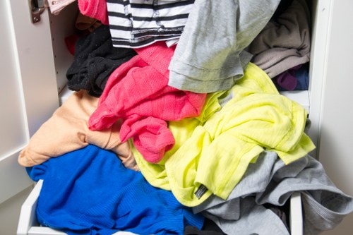 A close-up view of a white storage drawer filled with assorted clothes in various colours and fabrics, including a bright yellow garment, a blue piece, a pink item, and neutral tones such as grey and beige. The clothing appears wrinkled and casually piled, with some items partially spilling over the edges of the drawer. The background shows part of a white shelving unit or cabinet. The environment suggests an indoor setting, possibly a laundry room or storage area, relevant to household waste management services offered in the Yiewsley area. The image emphasizes the collection of textiles before disposal or recycling as part of rubbish removal services, with a focus on clothes management aspects.