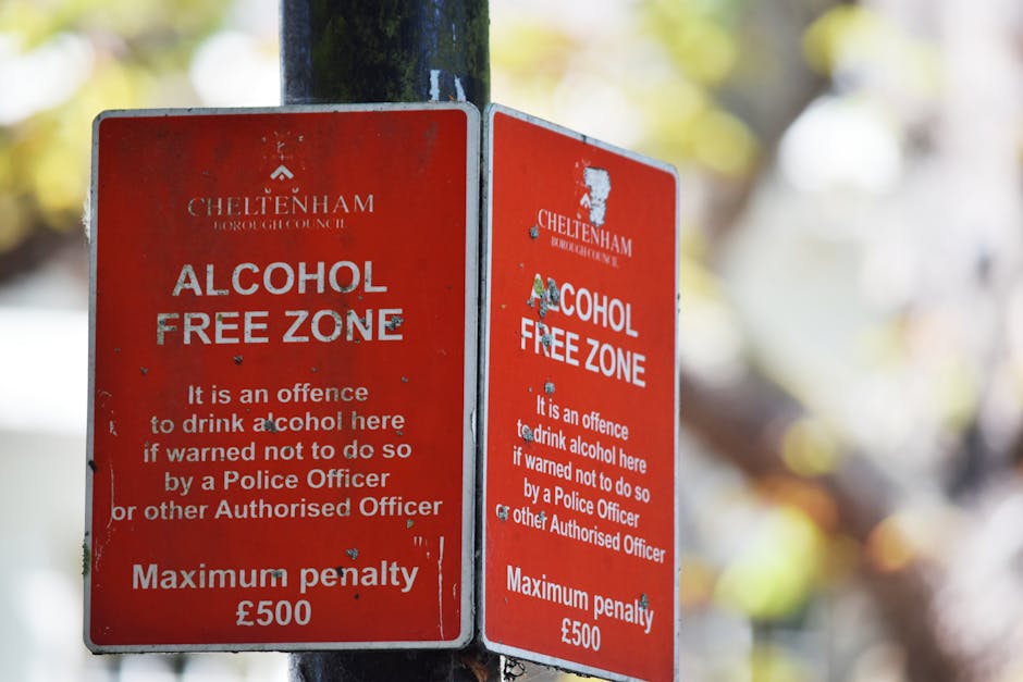 A pair of weathered red metal signs attached to a dark green metal pole, indicating a designated alcohol-free zone in Cheltenham. The signs feature white text warning that drinking alcohol in this are
